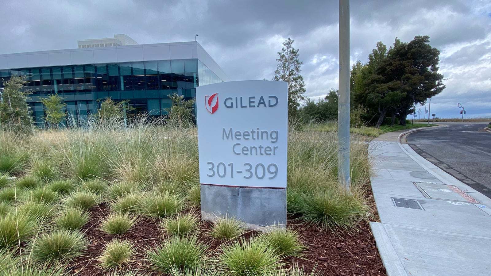 A white Gilead sign reading Meeting Center 301-309 stands among ornamental grasses in front of a glass-walled office building under a cloudy sky.