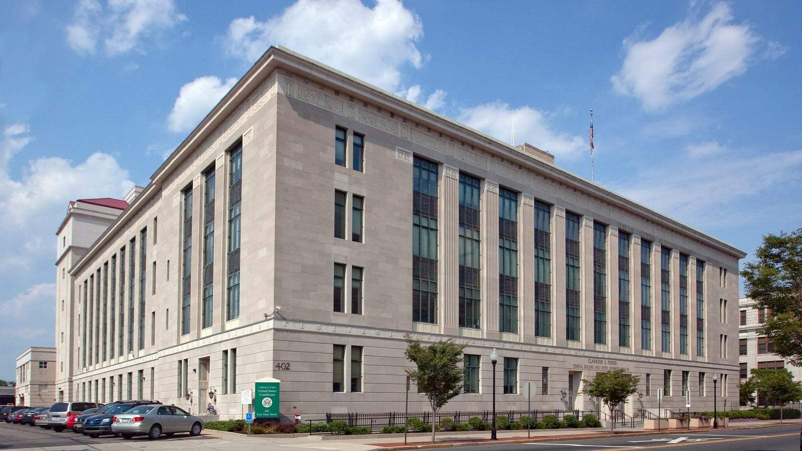 A four-story limestone federal courthouse with tall vertical windows on a street corner in Trenton, New Jersey, under a blue sky, with an American flag on the roof.