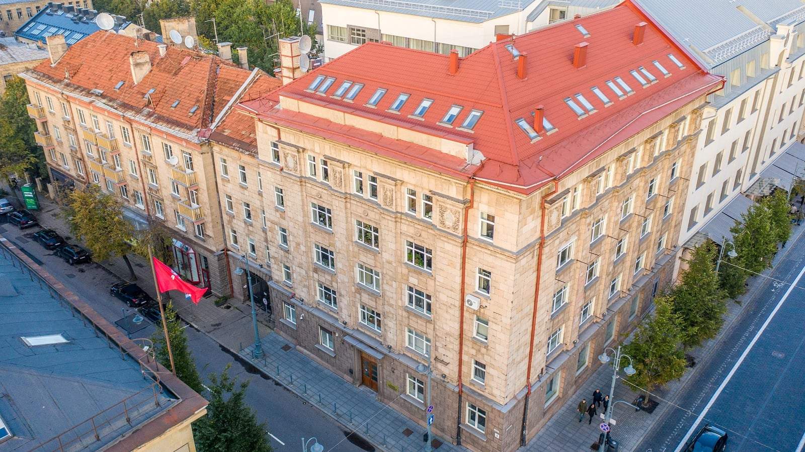 Aerial view of a pale stone multistory building with a red roof on a Vilnius street corner, with a red flag on a pole near the entrance.