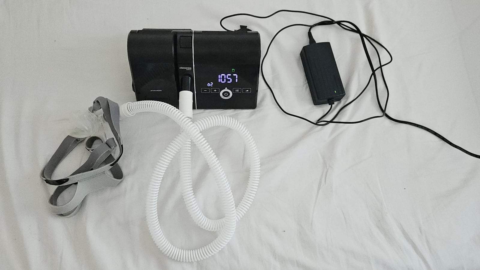 A CPAP machine on a white bed sheet, with a coiled white breathing hose, a gray mask headgear and a power adapter.