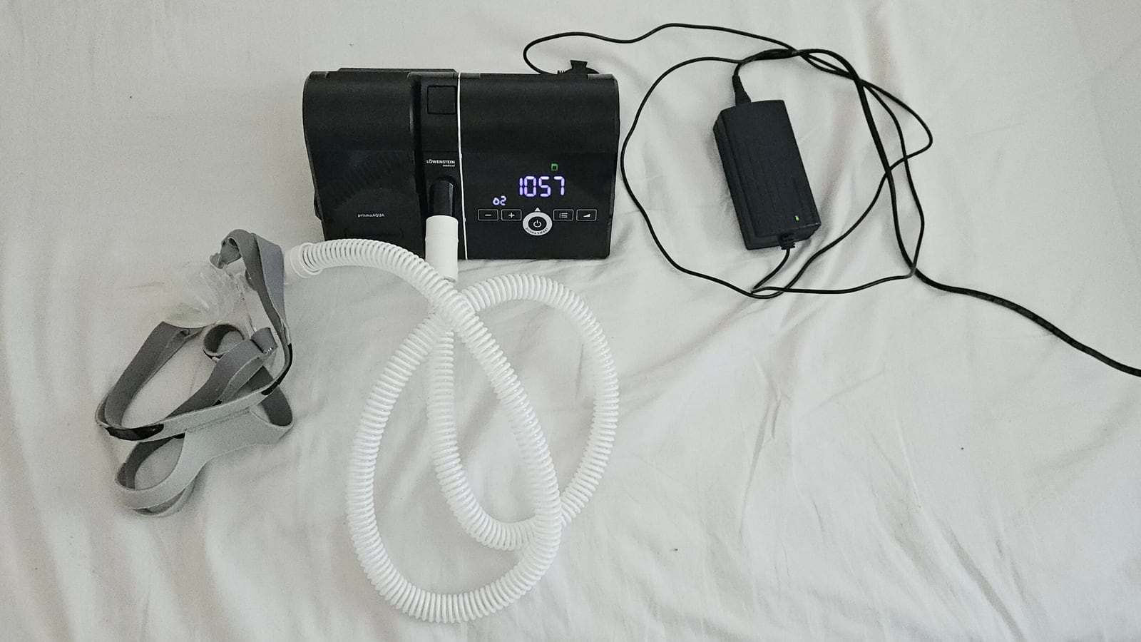 A CPAP machine on a white bed sheet, with a coiled white breathing hose, a gray mask headgear and a power adapter.