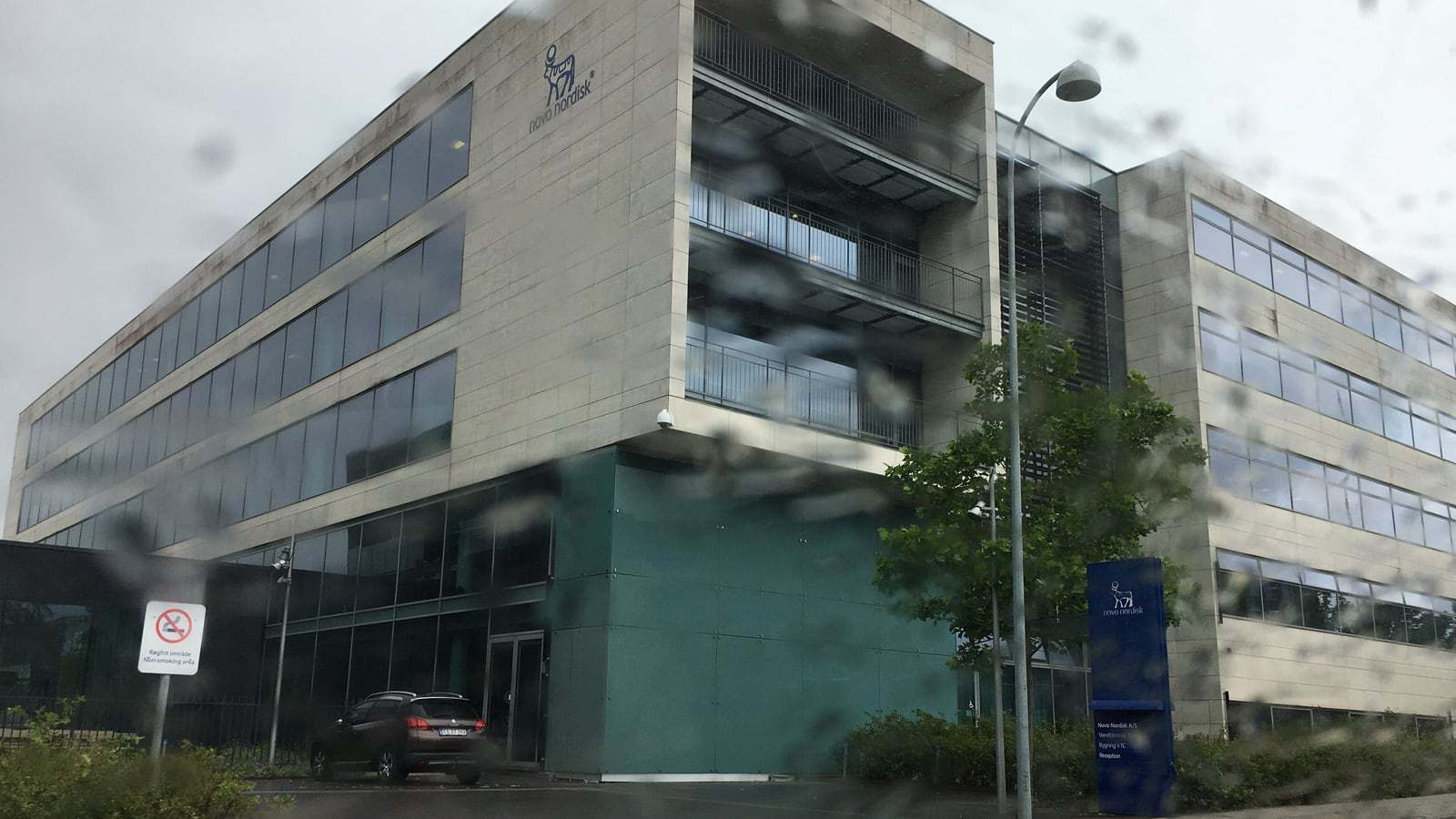 A multi-story Novo Nordisk office building with a blue glass facade and the company logo, photographed in rain in Søborg, Denmark.