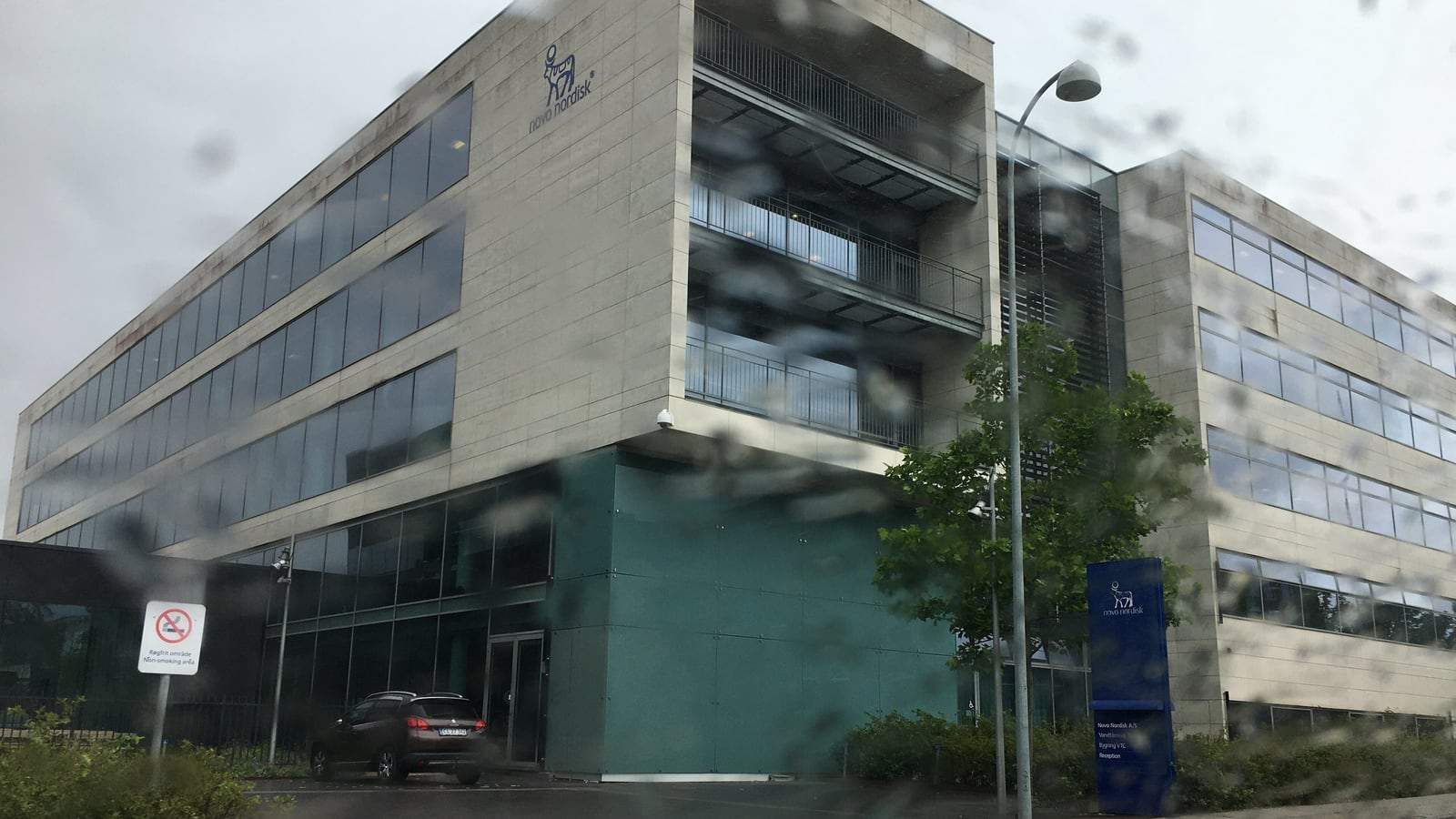 A multi-story Novo Nordisk office building with a blue glass facade and the company logo, photographed in rain in Søborg, Denmark.